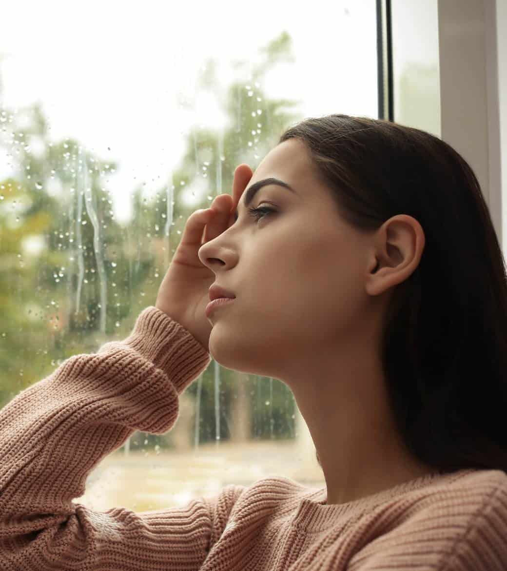 Woman sitting by a window on a rainy day, appearing overwhelmed.
